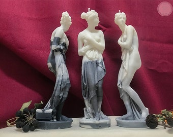 May include: Three white and gray sculpted candles in the shape of a woman standing with a draped cloth. The candles are on a white surface with a red background.