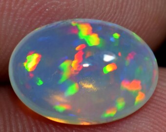 Può includere: Una gemma di opale traslucida, di forma ovale. L'opale mostra un colore di base bianco latte con vibranti lampi di rosso, verde e giallo, creando un effetto di gioco di colori. La gemma è liscia e lucidata, riflettendo la luce.