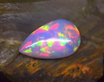 May include: A pear-shaped opal gemstone with a vibrant display of colors, including pink, green, and yellow, set against a dark, textured background. The gemstone's surface reflects light, creating a shimmering effect.
