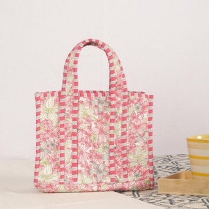 Peut inclure: Un sac fourre-tout &agrave; fleurs avec un motif de fleurs roses et blanches. Le sac a une forme rectangulaire avec une poign&eacute;e assortie et des rayures roses verticales. Le sac est fait d'un tissu matelass&eacute;.