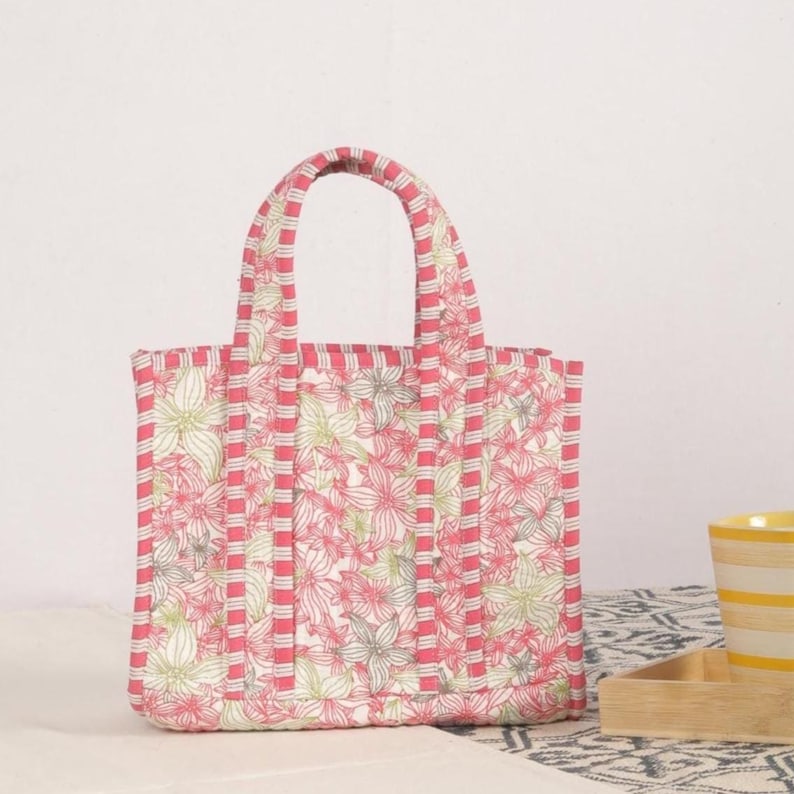 Peut inclure: Un sac fourre-tout &agrave; fleurs avec un motif de fleurs roses et blanches. Le sac a une forme rectangulaire avec une poign&eacute;e assortie et des rayures roses verticales. Le sac est fait d'un tissu matelass&eacute;.