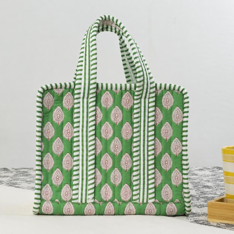 May include: Green tote bag with a repeating leaf pattern in white and red. The bag has white and green striped handles and trim. The bag is quilted and appears to be made of fabric.