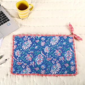 May include: A blue floral-patterned laptop sleeve with a red and white striped border. The sleeve features a quilted design with a repeating pattern of flowers in shades of blue, pink, and white. A yellow mug and a laptop are in the background.