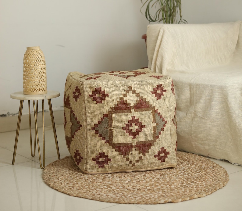Puede incluir: Un puf cuadrado estampado con un dise&ntilde;o geom&eacute;trico marr&oacute;n, beige y rojo. El puf est&aacute; sobre una alfombra de fibra natural.