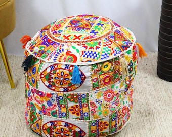 May include: A round, patchwork pouffe with a vibrant, multi-coloured design. The pouffe features floral and geometric patterns in red, yellow, blue, and green. It has decorative tassels in various colours and is likely used for seating or as a footrest.