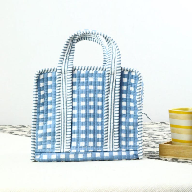 May include: A small, square tote bag with a blue and white gingham pattern. The bag has two handles and is trimmed with a striped fabric. The bag is sitting on a white surface.