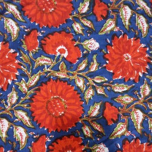 May include: Close-up of a fabric with a repeating floral pattern. Large, vibrant red flowers with detailed petals are scattered across a deep blue background. Green and white leaves and stems intertwine, creating a lively and colorful design.