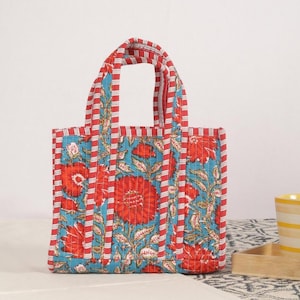 Peut inclure: Un sac fourre-tout matelass&eacute; avec un motif floral. Le sac pr&eacute;sente un design vibrant avec des fleurs rouges, un fond turquoise et des poign&eacute;es et bordures ray&eacute;es rouges et blanches. Le sac mesure environ 25 cm de haut.