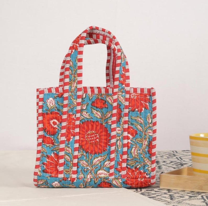 Peut inclure: Un sac fourre-tout matelass&eacute; avec un motif floral. Le sac pr&eacute;sente un design vibrant avec des fleurs rouges, un fond turquoise et des poign&eacute;es et bordures ray&eacute;es rouges et blanches. Le sac mesure environ 25 cm de haut.
