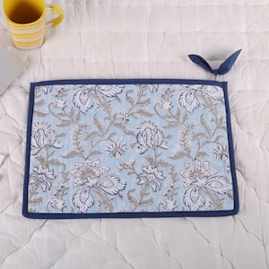 May include: A rectangular, floral-patterned placemat with a light blue background and white and gray flower designs. The placemat has a dark blue border and a loop for hanging. A yellow mug is visible in the background.