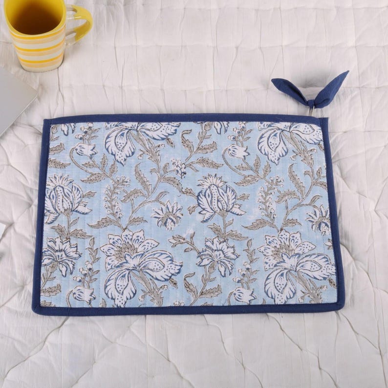 May include: A rectangular, floral-patterned placemat with a light blue background and white and gray flower designs. The placemat has a dark blue border and a loop for hanging. A yellow mug is visible in the background.