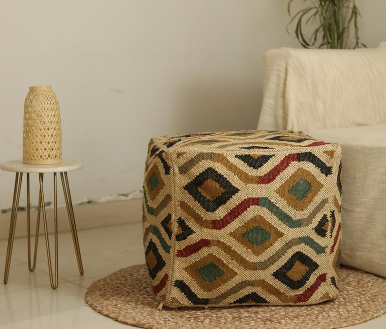 Puede incluir: Un puf cuadrado estampado con un dise&ntilde;o geom&eacute;trico en tonos marrones, negros, rojos y verdes. El puf est&aacute; hecho de yute tejido y tiene un aspecto natural y terroso.