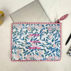 Könnte beinhalten: Eine weiße Laptoptasche mit einem Blumenmuster aus rosa Blüten und blauen Blättern. Die Tasche hat einen rosa-weiß gestreiften Rand und der Name "Jennifer" ist in Rosa aufgedruckt. Eine rosa-weiß gestreifte Stoffschlaufe ist befestigt.