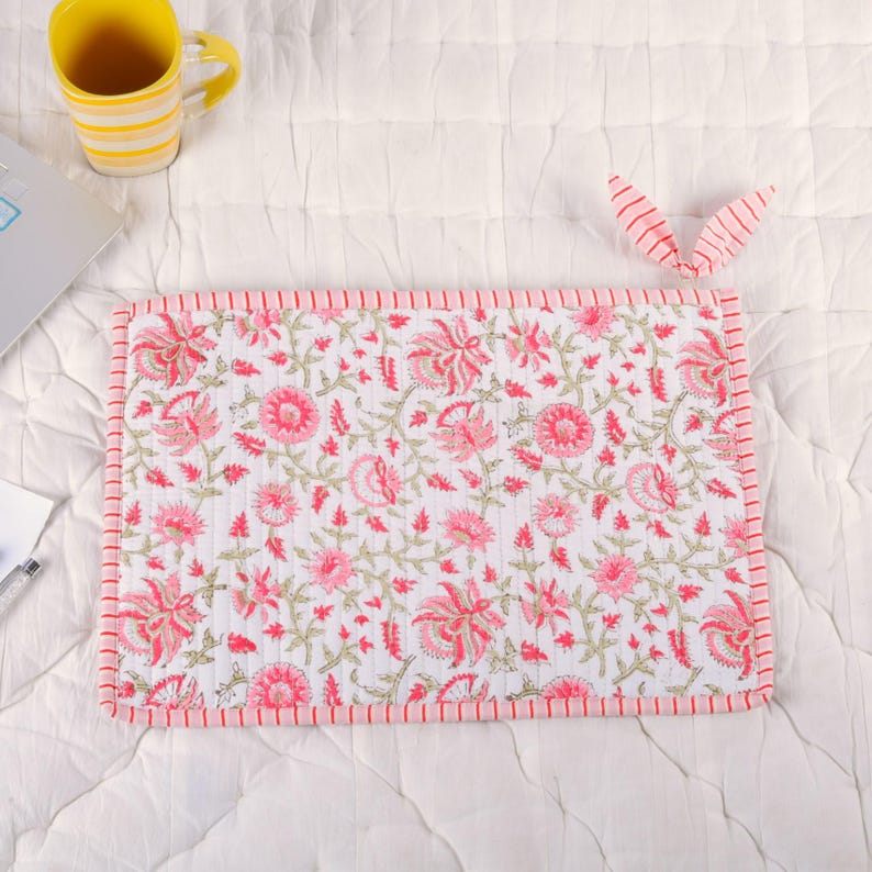 May include: A rectangular white and pink floral print laptop sleeve with a red striped border. The sleeve is surrounded by a white quilted surface, a yellow and white striped mug, and a pink striped bow.