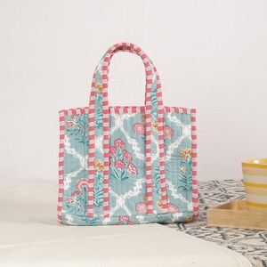 Peut inclure: Sac fourre-tout matelass&eacute; avec un motif floral dans les tons de turquoise, rose et jaune. Le sac a une forme rectangulaire avec deux anses et une bordure ray&eacute;e rose et blanche. Le sac est pos&eacute; sur une surface blanche.