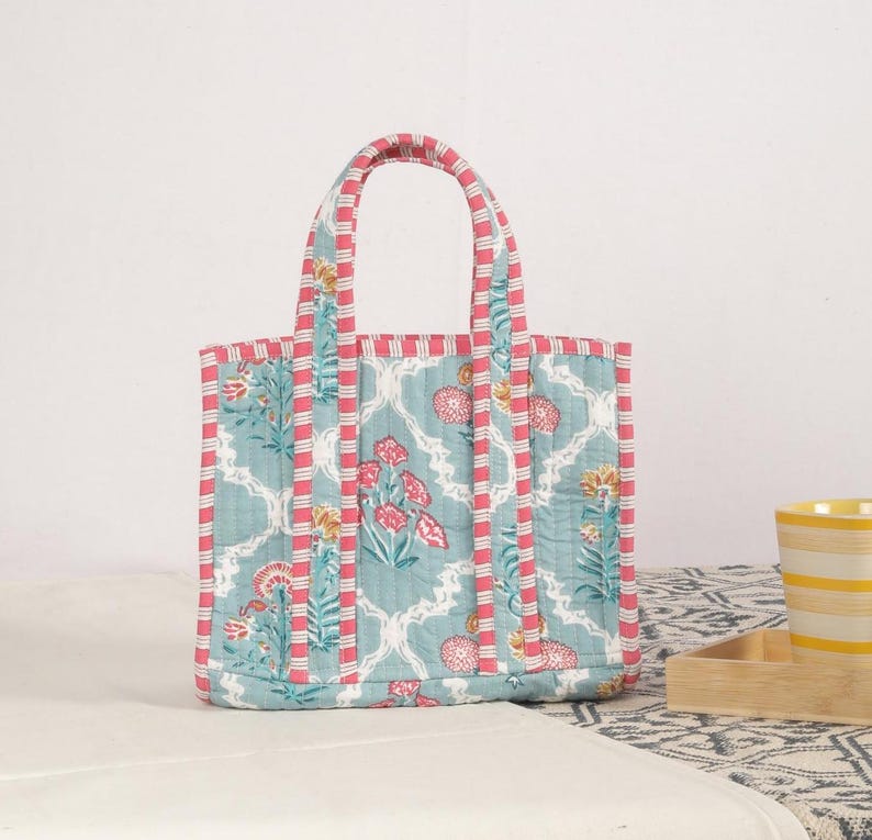 Peut inclure: Sac fourre-tout matelass&eacute; avec un motif floral dans les tons de turquoise, rose et jaune. Le sac a une forme rectangulaire avec deux anses et une bordure ray&eacute;e rose et blanche. Le sac est pos&eacute; sur une surface blanche.