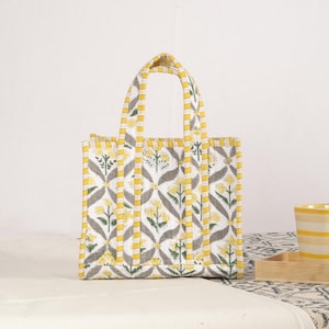 Peut inclure: Sac fourre-tout rectangulaire avec un motif floral jaune, gris et blanc. Le sac a deux poign&eacute;es et est fait d'un tissu matelass&eacute;. Le motif floral pr&eacute;sente des fleurs jaunes et des feuilles grises sur fond blanc.