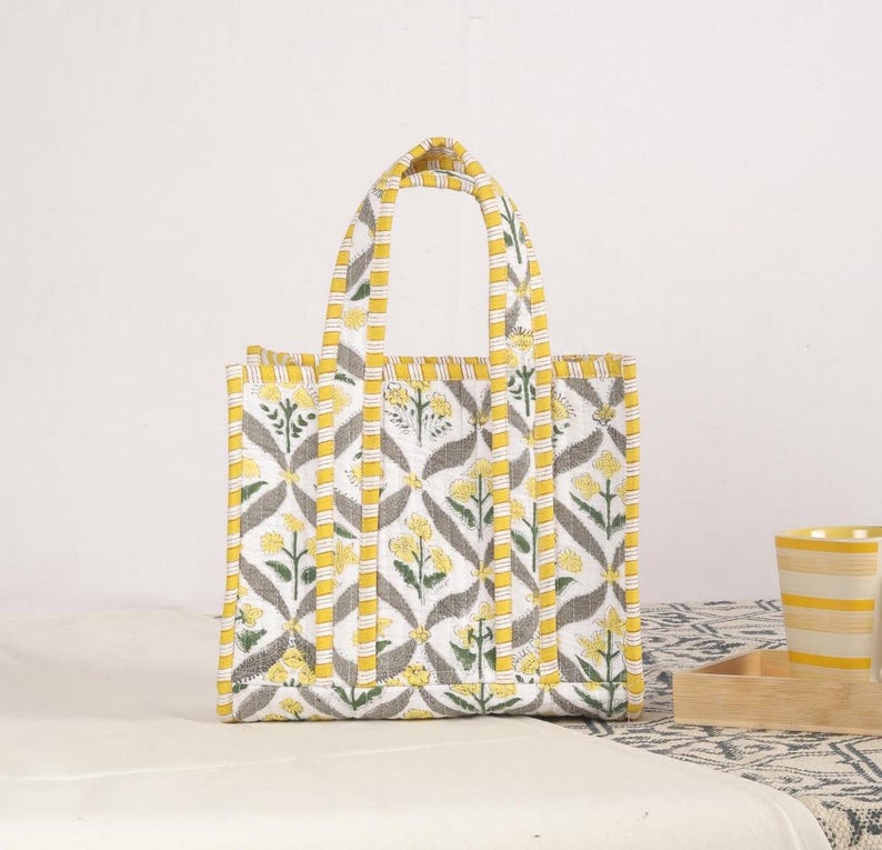 Peut inclure: Sac fourre-tout rectangulaire avec un motif floral jaune, gris et blanc. Le sac a deux poign&eacute;es et est fait d'un tissu matelass&eacute;. Le motif floral pr&eacute;sente des fleurs jaunes et des feuilles grises sur fond blanc.