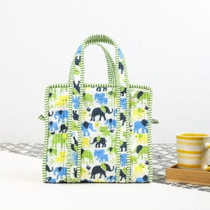 May include: A rectangular tote bag with a white background and a repeating pattern of blue, green, and yellow elephants. The bag has green striped handles and trim. A yellow mug with horizontal stripes sits on a wooden tray.