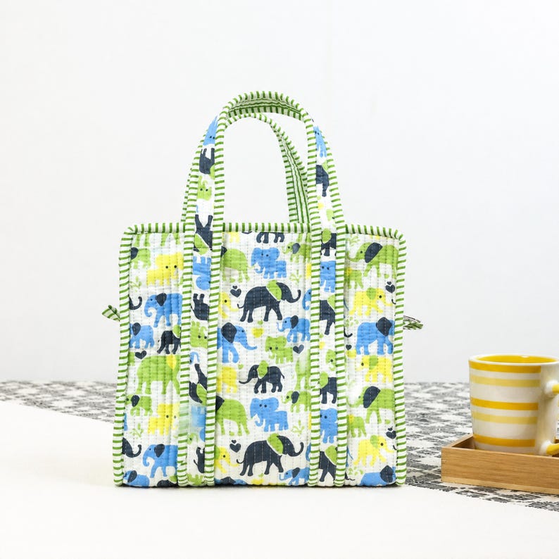 May include: A rectangular tote bag with a white background and a repeating pattern of blue, green, and yellow elephants. The bag has green striped handles and trim. A yellow mug with horizontal stripes sits on a wooden tray.