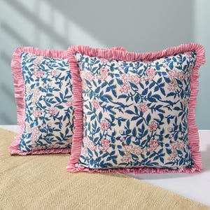 Puede incluir: Dos cojines cuadrados con fondo blanco, hojas azules y flores rosas. Los cojines tienen un ribete de volantes a rayas rosas y blancas. Los cojines están sobre una superficie beige.