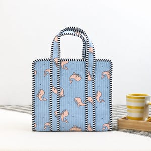 May include: A light blue tote bag with a repeating pattern of pink whales. The bag has black and white striped trim and handles. The bag is sitting on a white surface.