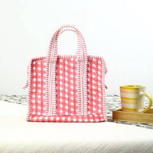 May include: A red and white gingham tote bag with a quilted design and matching handles. A yellow and white striped mug sits on a wooden tray, adding a touch of color to the scene.