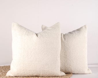 May include: Two white fluffy throw pillows with a subtle texture. The pillows are placed on a woven rug.