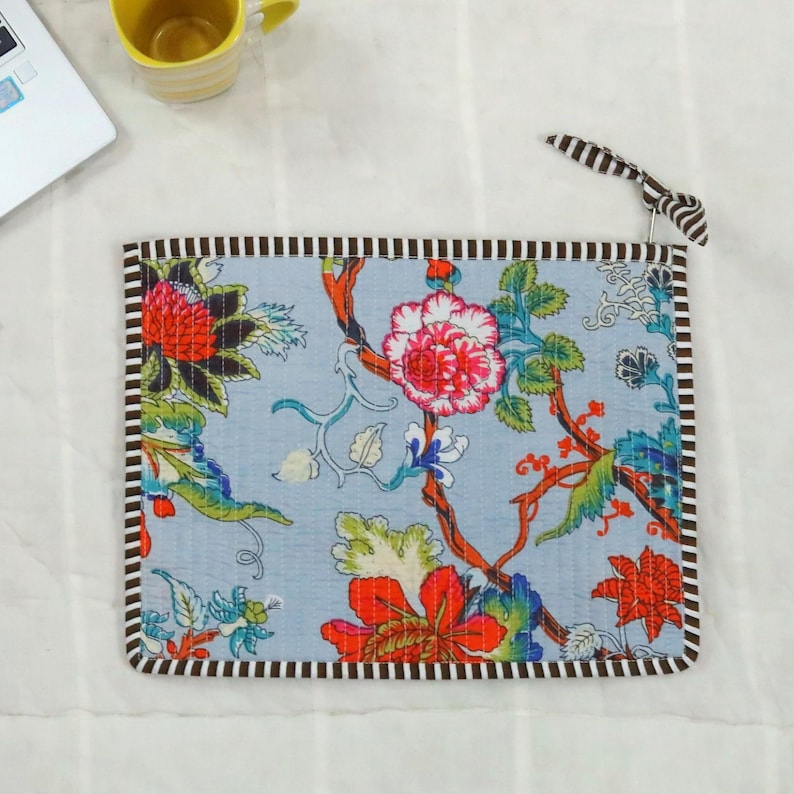 May include: A rectangular, light blue fabric item with a floral design in red, pink, and green. The edges are trimmed with a brown and white striped border. A black and white striped loop is attached to the top right corner.