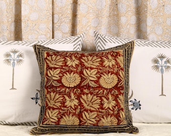 May include: A red and cream floral patterned throw pillow with a black border. The pillow is sitting on a white and gray patterned bedspread.