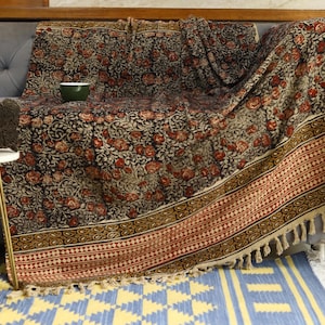 以下が含まれることがあります： グレーのソファに掛けられた、花柄のブランケット。ブランケットは、黒い背景に赤い花の繰り返し模様があり、装飾的なボーダーとフリンジが付いています。小さな緑色のボウルがブランケットの上にあります。