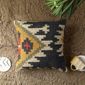 Puede incluir: Una almohada cuadrada con un patrón geométrico negro, blanco, amarillo, rojo y verde. La almohada está hecha de un tejido tejido y tiene una superficie texturizada.