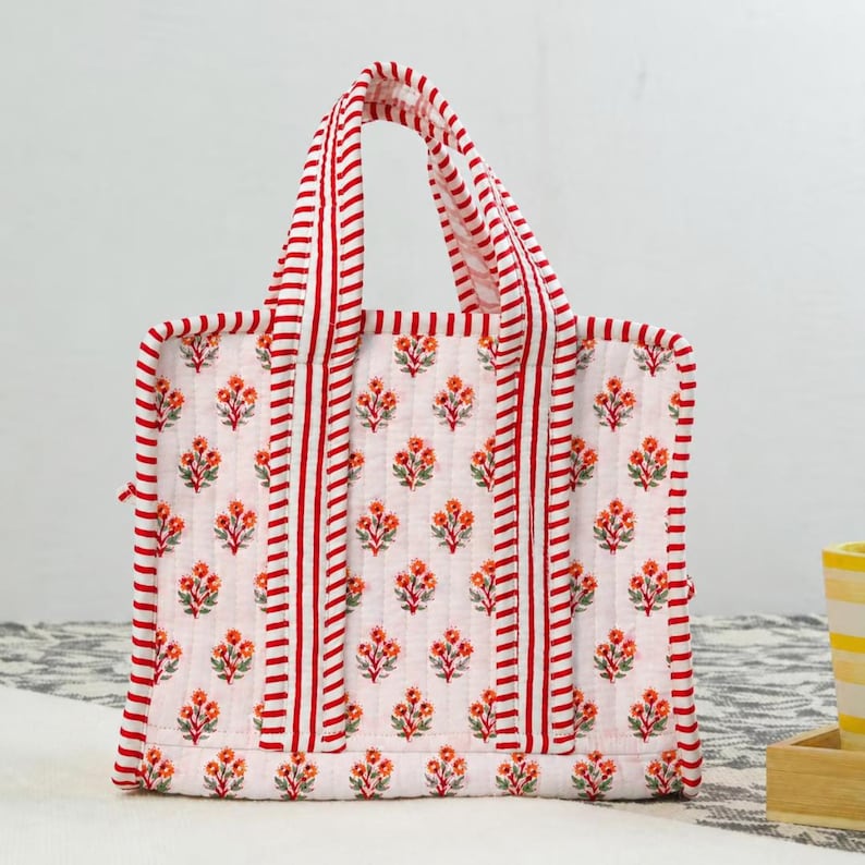 May include: A white tote bag with a floral pattern and red stripes. The bag features a repeating pattern of orange flowers and green leaves. The handles and edges are trimmed with red stripes.