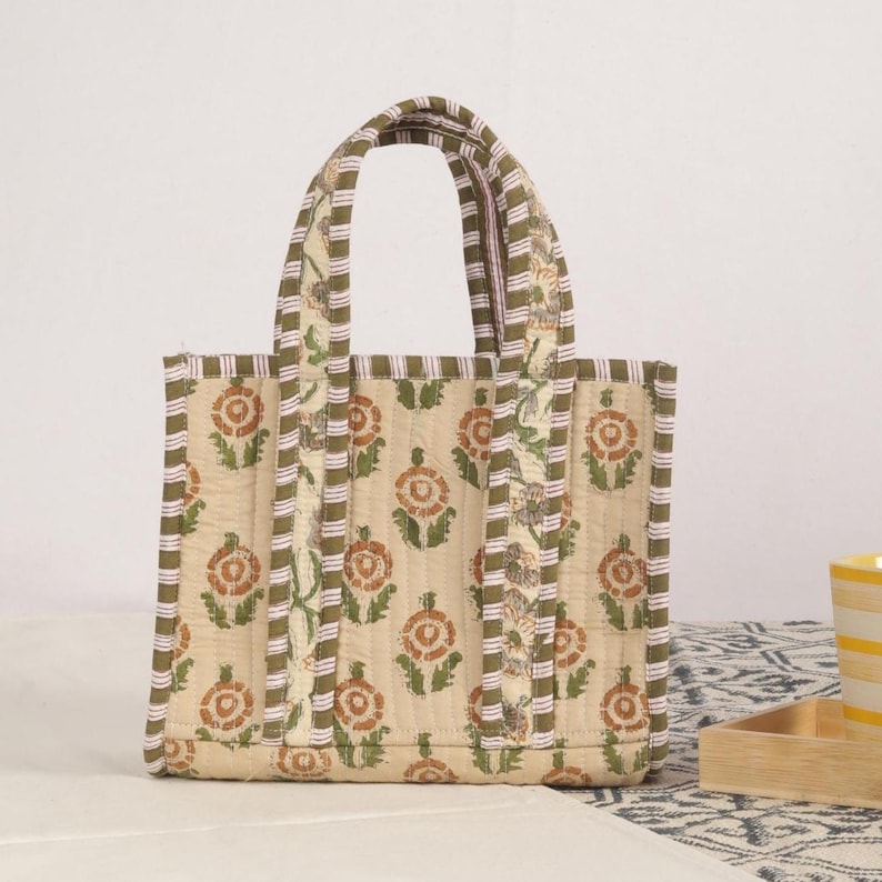 Peut inclure: Sac fourre-tout matelass&eacute; beige avec un motif floral orange et vert. Le sac a deux poign&eacute;es et est bord&eacute; d'un tissu ray&eacute; vert et blanc. Le sac mesure environ 25 cm de large et 20 cm de haut.