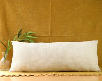 May include: A white rectangular lumbar pillow with a textured linen fabric. The pillow is lying on a gold fringed throw rug.