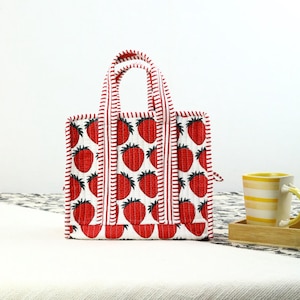 May include: A small, square tote bag with a white background and a pattern of red strawberries. The bag has red and white striped handles and trim. The bag is sitting on a white surface.