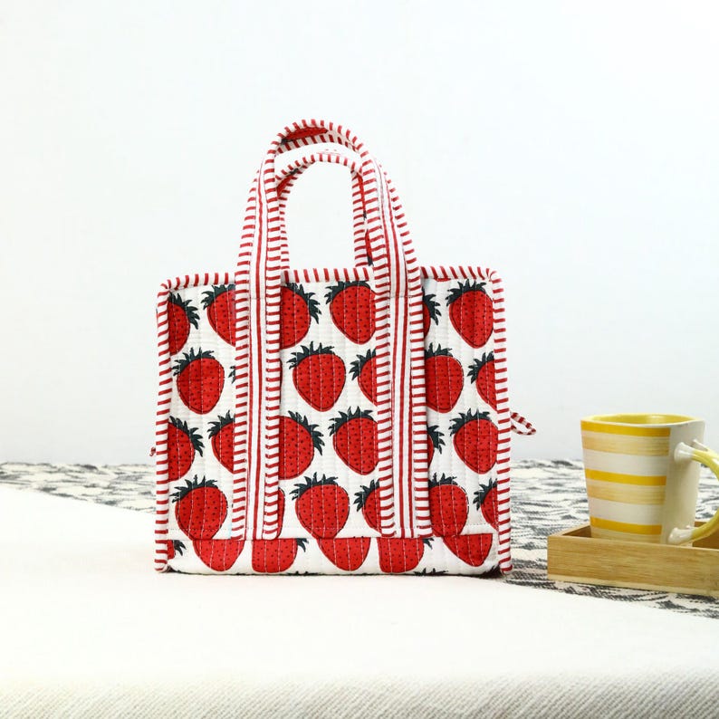 May include: A small, square tote bag with a white background and a pattern of red strawberries. The bag has red and white striped handles and trim. The bag is sitting on a white surface.