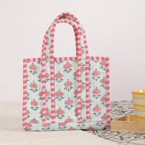 Könnte beinhalten: Eine gesteppte Tragetasche mit floralem Muster. Die Tasche hat einen hellblauen Hintergrund mit rosa Lotusblüten-Designs. Die Kanten und Griffe sind mit rosa und weiß gestreiftem Stoff verziert. Die Tasche ist mit zwei Griffen ausgestattet.