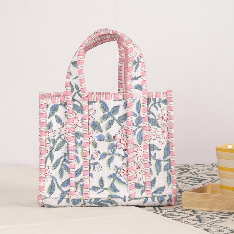 Peut inclure: Un petit sac fourre-tout matelass&eacute; avec un motif floral. Le sac pr&eacute;sente un fond blanc avec des fleurs roses et des feuilles bleues. Une bordure ray&eacute;e rose et blanche borde le sac et les poign&eacute;es. Le sac est pos&eacute; sur une surface &agrave; motifs.