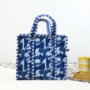 May include: A blue tote bag with a repeating pattern of elephants, giraffes, and lions. The bag has a quilted texture and white and blue striped handles and trim. The bag is rectangular with a square base.