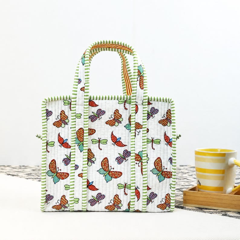 May include: A white tote bag with a colorful butterfly and dragonfly pattern. The bag has green trim and handles with a striped design. The bag is sitting on a white surface.