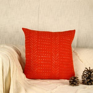 May include: A red throw pillow with a white geometric pattern. The pillow is on a white couch with two pine cones.