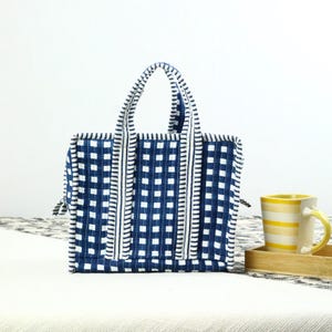 May include: A blue and white checkered tote bag with matching handles. The bag has a rectangular shape and is made of a quilted fabric. A yellow and white striped mug sits on a wooden tray next to the bag.