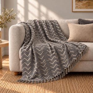 May include: A charcoal gray throw blanket featuring a repeating white chevron pattern and decorative fringe along the bottom edge. The blanket is draped over the arm and seat of a light-colored sofa, showcasing its textured fabric.
