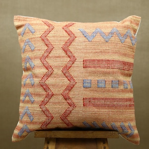 May include: Square decorative pillow with a woven texture. The pillow features a beige background with red and blue geometric patterns, including zigzags and horizontal lines. The pillow is placed on a wooden stool.