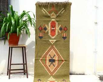 May include: Olive green woven rug with geometric patterns in red, tan, and black. The rug features a diamond and rectangular design with fringe at the top. The rug is displayed vertically.