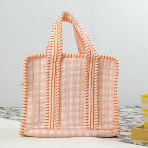 Könnte beinhalten: Eine gesteppte Tragetasche mit Pfirsich- und weißem Vichy-Muster. Die Tasche hat orange gestreifte Griffe und Besätze. Die Tasche hat eine kleine, quadratische Form und eignet sich zum Tragen kleiner Gegenstände. Die Tasche ist aus weichem, strukturiertem Stoff.