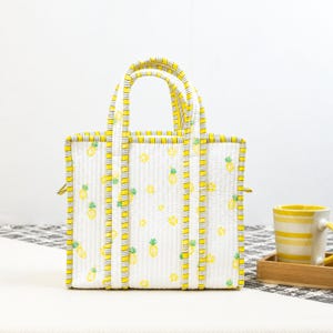 May include: White tote bag with a yellow pineapple pattern and yellow trim. The bag has two handles and a rectangular shape. A yellow and white striped mug sits on a wooden tray.