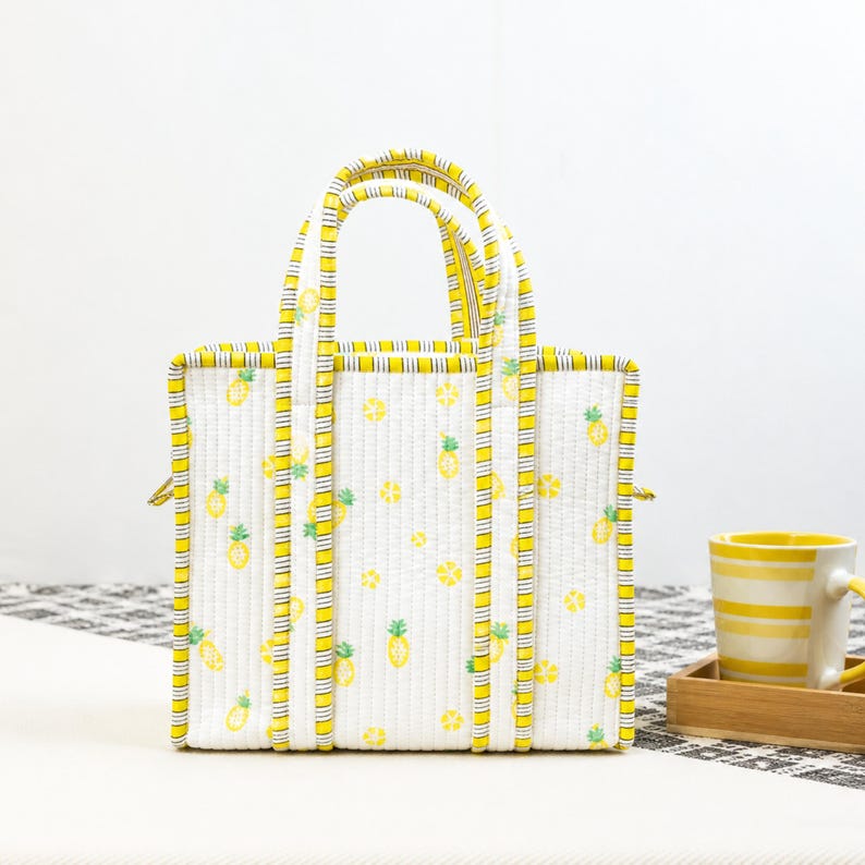 May include: White tote bag with a yellow pineapple pattern and yellow trim. The bag has two handles and a rectangular shape. A yellow and white striped mug sits on a wooden tray.