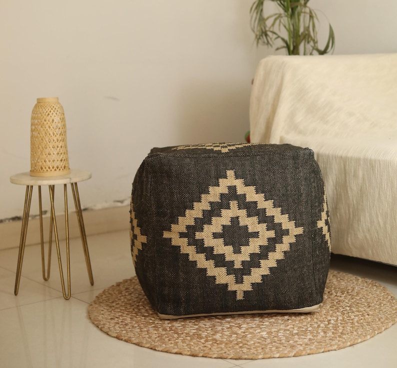 Puede incluir: Un puf cuadrado negro y beige con un dise&ntilde;o geom&eacute;trico. El puf est&aacute; sobre una alfombra beige.